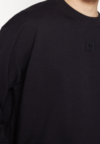 Sudadera negra hecha de algodón, con un cuello redondo, mangas largas y una pequeña etiqueta negra con el logo en el pecho. Textura suave.