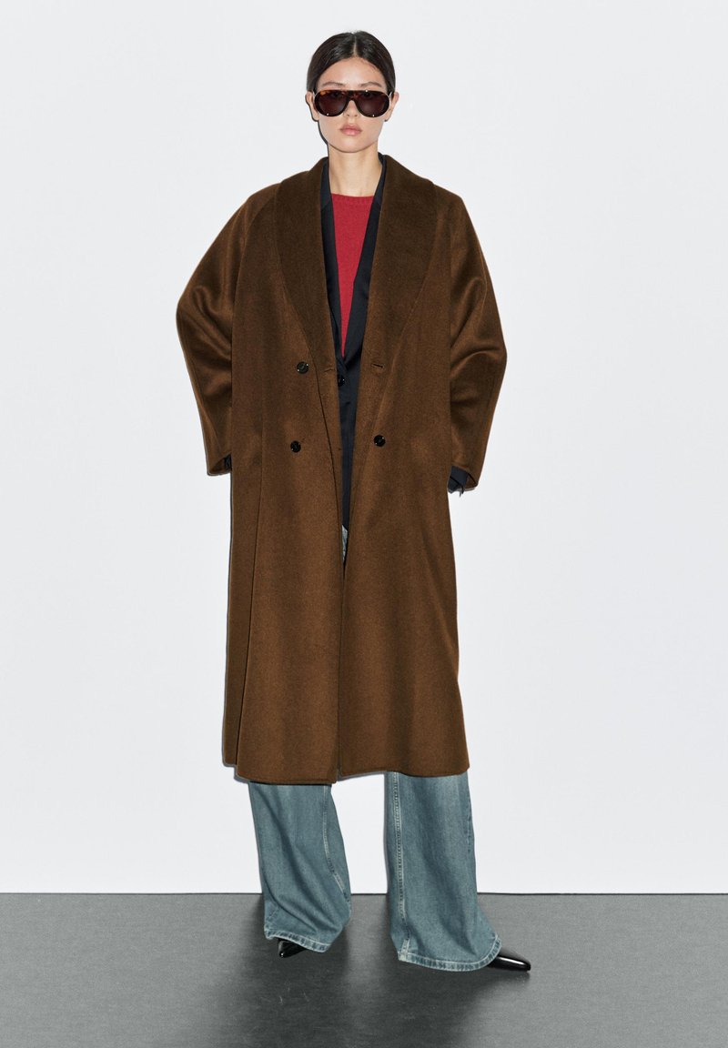 Brun dubbelknäppt kappa gjord av ett mjukt, texturerat tyg, matchad med en röd topp och vida ljusblå jeans. Tillbehör inkluderar mörka solglasögon.