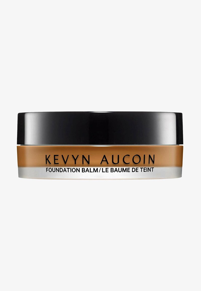 Kevyn Aucoin Foundation Balm in einem runden, schwarzen und klaren Behälter. Das Produkt hat eine warme Tan-Farbe mit glatter Textur.