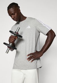 Pilkas sportinis marškinėliai su baltais "adidas" dryželiais ant rankovių, laikomi asmens, laikančio juodą ir sidabrinį svarmenį, prieš šviesų foną.