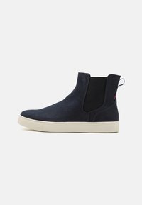 Marineblå ankelstøvletter i suede med elastiske sidepaneler og en struktureret beige gummisål. Har en strop og et minimalistisk design.