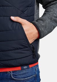 Ruka v zipové kapse námořnického prošívaného vesty, která je oblečená přes šedý pletený rukáv a červený svetr, doplněná modrými džíny.