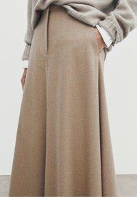 Jupe longue et fluide en beige fabriquée dans un tissu doux, avec une taille haute, une poche latérale et une silhouette élégante. L'ourlet arrive près du sol.