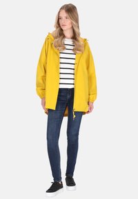 Sárga vízálló kabát kapucnival, kényelmes szabással és elasztikus mandzsettákkal, fekete-fehér csíkos ing és kék farmer fölött viselve.