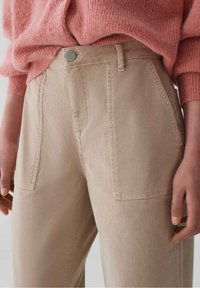 Pantalon en velours côtelé beige avec deux poches avant et une fermeture à bouton métallique. Associé à un pull rose texturé, ample.