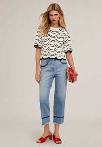 Top de punto de rayas onduladas en blanco y negro, jeans cortos de color azul claro con dobladillo deshilachado y sandalias de tacón cuadrado rojas. Sosteniendo un bolso pequeño rojo.