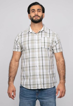 Homme aux cheveux foncés et barbe portant une chemise à manches courtes beige et blanche à carreaux boutonnée et un jean bleu, debout devant un fond gris.