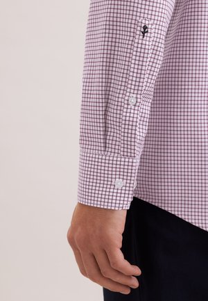 Hand van een man naast een lange mouw met een witte en paarse geruite boord, met witte knopen en een subtiel geborduurd logo.
