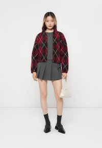 Rød og svart argyle-mønstret cropped jakke, grå tettsittende topp, grå plisserte skjørt, svarte sko og hvit veske. Glatte teksturer gjennomgående.