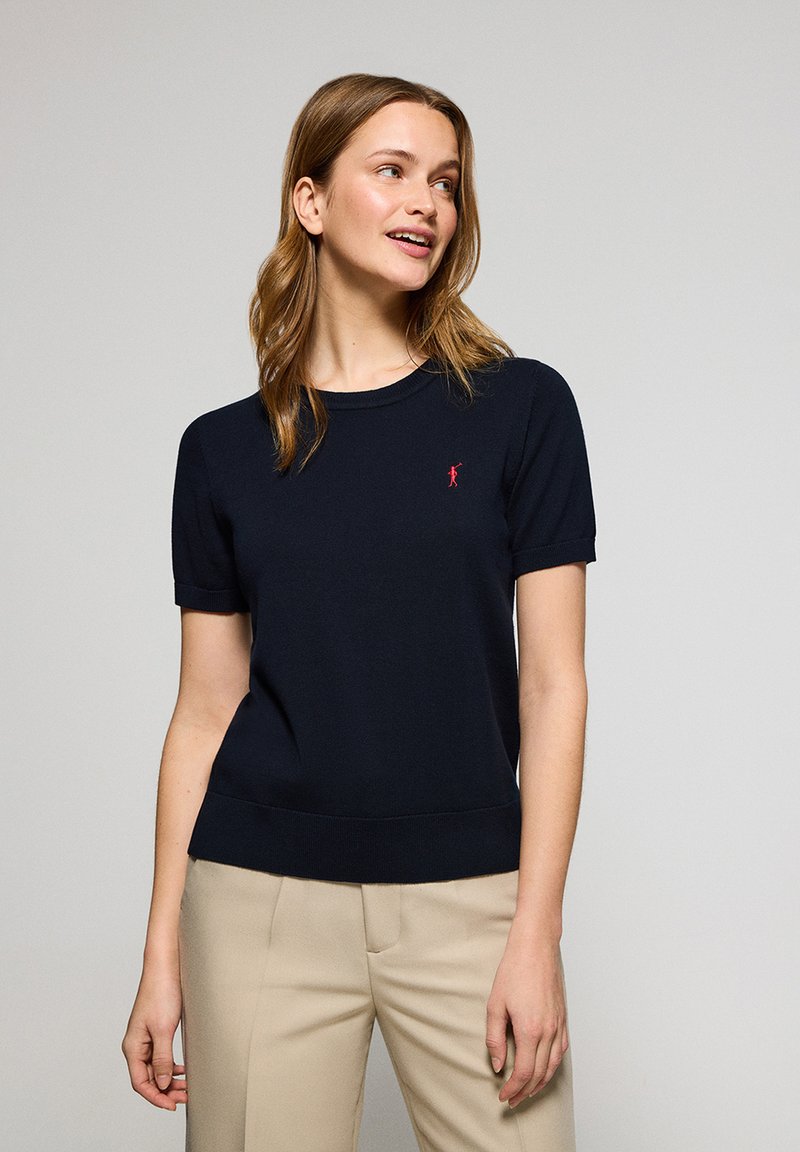 Granatowy wełniany sweter z krótkimi rękawami, okrągłym dekoltem, ściągaczem u dołu i małym czerwonym emblematem na lewej piersi.