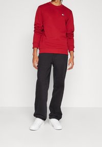 Sudadera roja de manga larga con puños acanalados, combinada con jeans negros. El modelo lleva zapatillas blancas. La sudadera presenta un pequeño logo.