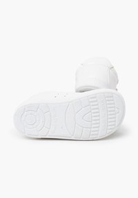 Biały dziecięcy tenisówka z fakturowaną gumową podeszwą, zaokrąglonym noskiem i przeszyciami wokół kostki. Na podeszwie wybite „Wyprodukowano w Hiszpanii”.