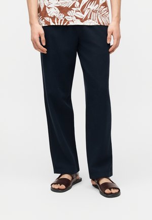 Osoba ubrana w czarne spodnie o prostych nogawkach, brązowe sandały z odkrytymi palcami oraz brązową koszulę w tropikalny wzór liści, stojąca na jednolitym tle.