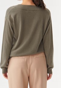 Pull vert olive surdimensionné avec une texture douce et des coutures d'épaule tombantes, associé à un pantalon tailleur rose pâle avec des poches arrière.