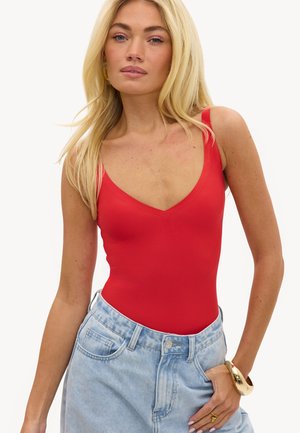 Femme blonde portant un haut rouge sans manches rentré dans un jean taille haute bleu clair, accessoirisée par un bracelet et une bague en or.
