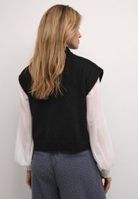 Black knitted sleeveless top with a rounded neckline, layered over a white textured blouse. Grey polka-dot trousers in the background.