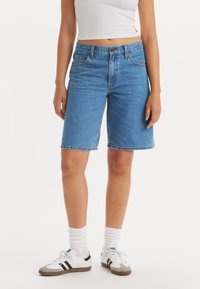 Denim bermuda kratke hlače v srednji modri barvi, s straight cut obliko, klasičnim pet-imenskim dizajnom in šivanjem na robovih. Nosijo se z belimi športnimi čevlji in nogavicami.