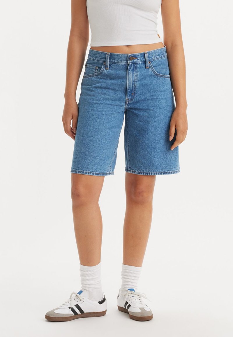 Denim bermudashorts i medelblått, med rak skärning, klassisk fem-fickorsdesign och synlig söm. Bärs med vita sportskor och strumpor.