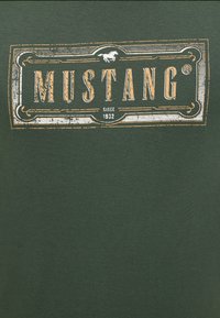 Tissu vert foncé avec une étiquette rectangulaire usée portant "MUSTANG" en grandes lettres, un petit icône de cheval au-dessus et "depuis 1932" en dessous.