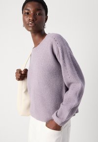 Lavendel strikket genser med rund hals, ribbestruktur og sidespalter. Kombinert med en hvit veske og stylet med lyse bukser.