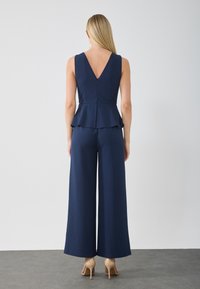 Combinaison peplum bleu marine avec un dos en V, design à jambes larges et texture lisse. Fermée par une fermeture éclair à l'arrière ; présente un détail peplum superposé.