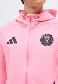 Osoba oblečená v růžové bundě na zip s kapucí, na hrudi s černými logy Adidas a Club Internacional de Fútbol Miami.