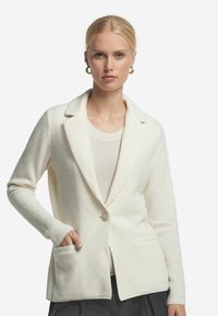 Weißes strukturiertes Blazer mit Schalkragen, Knopfverschluss und zwei Fronttaschen. Darunter ein hellcremes Oberteil.