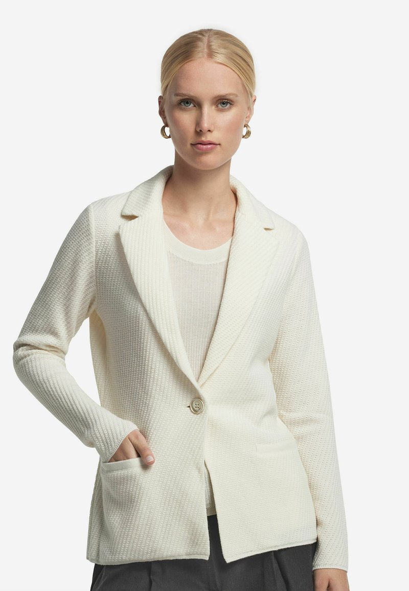 Weißes strukturiertes Blazer mit Schalkragen, Knopfverschluss und zwei Fronttaschen. Darunter ein hellcremes Oberteil.
