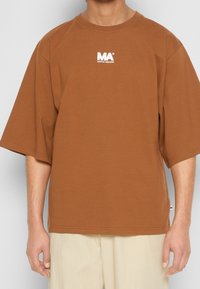 Camiseta marrón de gran tamaño, fabricada en algodón. Presenta un logotipo blanco en el pecho. Mangas cortas y sueltas con cuello redondo. Tejido texturizado.