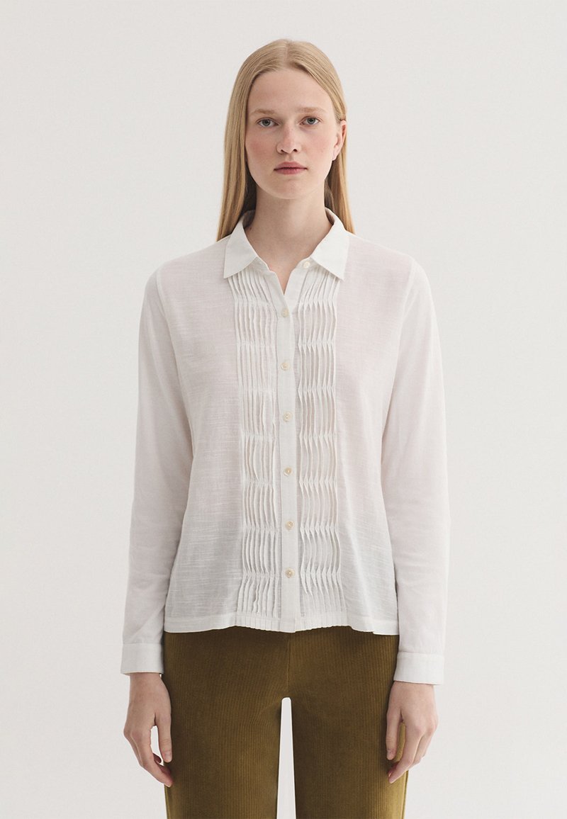 Femme aux longs cheveux blonds portant une chemise blanche boutonnée avec des détails plissés et un pantalon vert olive, debout devant un fond uni.
