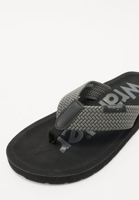 Svart flip-flop sandal med et vevd grått stropp og en teksturert såle. Har logo trykt på fotsengen og er laget av slitesterkt gummi materiale.