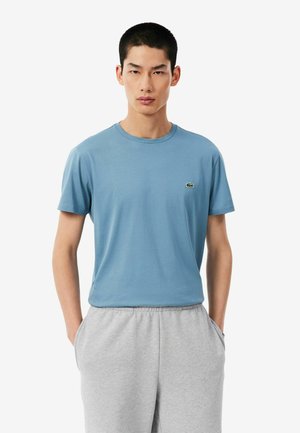 Jonge man die een lichtblauw T-shirt met korte mouwen en lichtgrijze joggingbroek draagt, staat met zijn handen in zijn zakken.