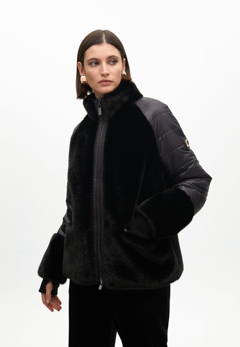 Black faux fur and quilted nylon jacket with a high collar, zip closure, and wide sleeves. Features pocket on the left arm.