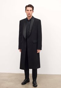 Palton lung negru cu rever din satin, ajustat, purtat peste o cămașă neagră cu nasturi și pantaloni, completat cu pantofi eleganți negri.