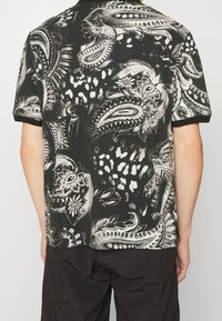 Koszula krótki rękaw w kolorze czarnym z białymi wzorami paisley i abstrakcyjnymi; miękka tekstura; posiada standardowy kołnierzyk oraz kontrastowe czarne wykończenia.