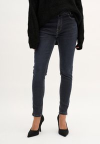 Jeans skinny azul oscuro de cintura alta, con una cremallera en el tobillo, combinados con tacones negros de punta y un suéter negro oversize.