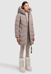 Polstret, taupe-farvet frakke med et chevron-design, der har en stor hætte og lynlåslukning. Sammen med beige leggings og hvide, pelsforede støvler.