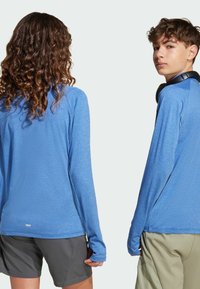 Modré športové tričká s dlhým rukávom vyrobené z ľahkej tkaniny, s voľným strihom a otvormi na palce, spárované so šedými šortkami.