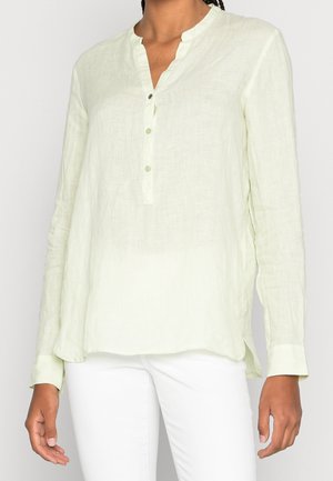 Kvinde iført en lysegrøn bluse med lange ærmer og kort knapstykke, kombineret med hvide bukser, stående mod en ensfarvet baggrund.