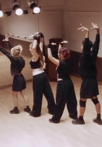 Čtyři tanečníci v zkušebně, oblečeni v černém s různými styly, předvádějí dynamické pohyby pažemi. Teplé osvětlení zvýrazňuje jejich pózy.