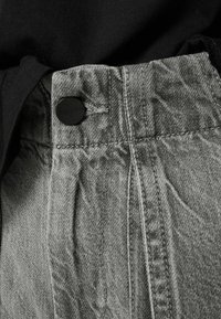 Grijze denim broek met een gestructureerd oppervlak, voorzien van een zwarte knoopsluiting en zichtbare stiksels langs de tailleband en naden.