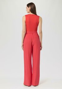 Czerwona bezrękawnik zestawiona z spodniami o wysokim stanie i szerokich nogawkach w pasującym odcieniu. Gładka tekstura z subtelnym tylnym detalem i ukrytymi zapięciami.