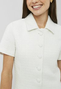 Robe blanche texturée avec un col, des manches courtes et quatre grands boutons sur le devant.