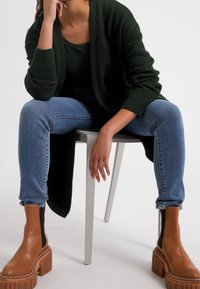 Grön stickad cardigan, mörkblå jeans och bruna klumpiga klackstövlar. Modell sitter på en metallstol med en neutral bakgrund.