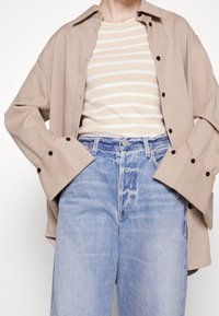 Beżowa koszula zapinana na guziki z czarnymi guzikami, noszona na jasną koszulkę w paski, w zestawieniu z jasnoniebieskimi dżinsami z lekko wyblakłym wykończeniem.