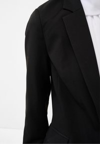 Černý blejzr s přizpůsobeným střihem, strukturovanými ramenními vycpávkami a hladkým povrchem. Obsahuje klopy a boční kapsy.