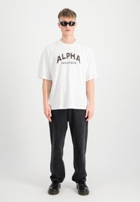 Biela bavlnená tričko s logom "ALPHA INDUSTRIES" v čiernej a oranžovej farbe, skombinované s voľnými čiernymi džínsami a čiernymi topánkami.