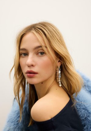 Orecchini pendenti tono argento con sfaccettature rotonde che riflettono la luce. Il modello ha capelli biondi fino alle spalle; indossa una maglia blu scuro con scollo a barca.