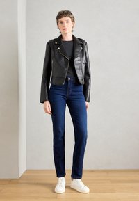 Črna umetna usnjena kratka jakna s srebrnimi zadrgami, kombinirana z visokim pasom temno modrimi kavbojkami in belimi športnimi čevlji na leseni tla.