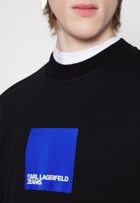 Felpa nera con un grafico a quadrato blu e la scritta "KARL LAGERFELD JEANS". Tessuto liscio con colletto a girocollo e uno strato interno a contrasto.